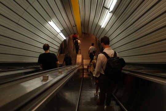 Travelers on escalators descend into a busy transit station in the Eternal City. The metallic, ribbed tunnel creates a contemporary backdrop for the moving public, highlighting the pulse of city exist