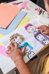 Child working on a fun art project with colorful paper hair glued onto cartoon faces. Close-up view...