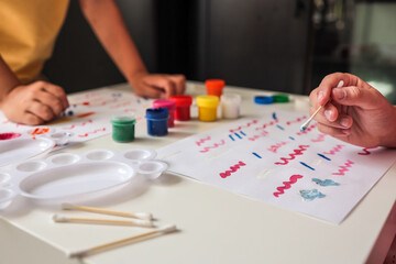 Fototapeta premium Kids using cotton swabs to paint colorful abstract lines on paper. Art supplies, paint jars, and palettes are on the white table. Hands in action during a creative DIY art session