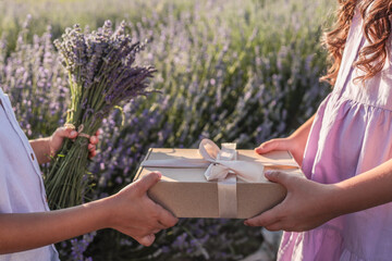 Close-up of two children exchanging a kraft gift box with a ribbon and a lavender bouquet in a...