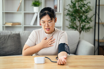 An Asian woman uses a portable blood pressure monitor in her living room. Health awareness concept.