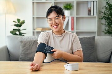 An Asian woman uses a portable blood pressure monitor in her living room. Health awareness concept.