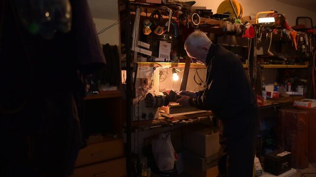 Grandpa sharpens an axe in his workshop. In a cozy workshop filled with tools, an elderly craftsman carefully adjusts a vintage sewing machine, his dedication illuminated by a warm lamp.