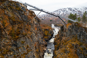 Impressive Voringsfossen footbridge spans over deep gorge with winding river against snowy mountains. Dramatic natural landscape in Norway region