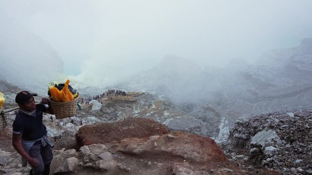 Miner carrying extremely heavy sulfur load out of Kawah Ijen volcano crater during the day in East Java, Indonesia, wide shot