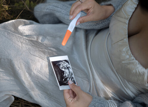 Pregnant woman in park or wild nature holding belly, ultrasound image and positive pregnancy test. Girl in first trimester sitting in grass wearing dress.