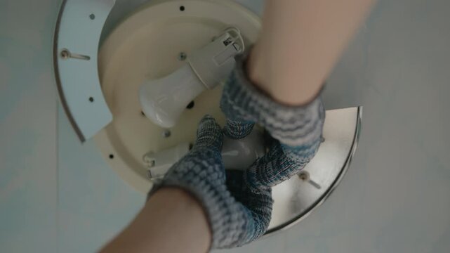 Close-up of unrecognizable electrician in protective gloves carefully replacing light bulb in ceiling light fixture. Concept of working with lighting appliances in home. Shooting in slow motion.