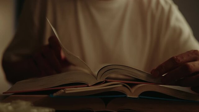 Close-up of male hands turning page of open book, resting on stack of books at table in dark living room, fostering warm and cozy atmosphere perfect for reading and studying. Shooting in slow motion.