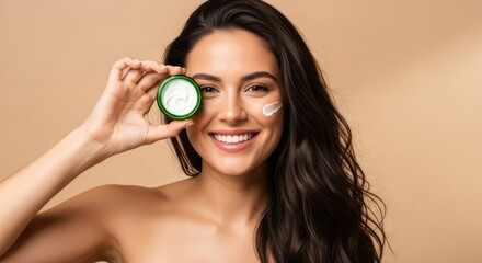 A beautiful young woman smiles while holding a jar of skincare cream to her cheek