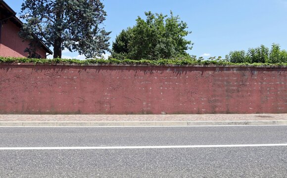 Weathered red plaster boundary wall at the roadside. Khy and trees on behind,sidewalk and street in front. Background for copy space.