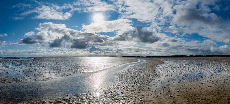 Panorama &uuml;ber das glitzernde Wattenmeer der Nordsee bei Ebbe, bzw. Niedrigwasser nit der leicht bedeckten Sonne &uuml;ber dem Horizont und blauem Himmel bei aufgelockerter Bew&ouml;lkung