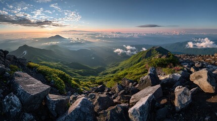 Naklejka premium Panoramic mountain vista at dawn. Vast landscape of jagged peaks, valleys, and clouds. Lush green slopes and scattered rocks. Sunrise hues paint the sky