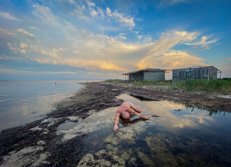 Nude figure in water, cloudy sky, rustic shore, black and white.
