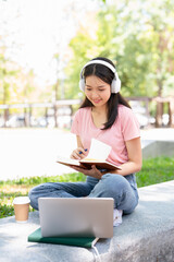 Obraz premium Smiling young asian woman in wireless headphones sitting outdoor working on laptop computer and writing or taking notes in paper notebook, holding pen in hand.