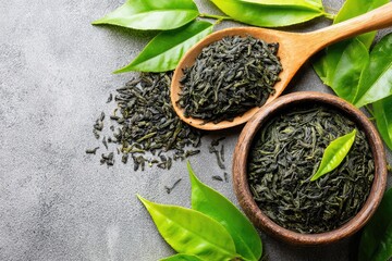 Dried green tea leaves in wooden bowl and spoon, fresh green leaves on gray surface