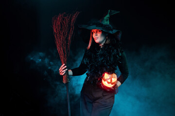 Young woman wearing halloween witch costume on black background with magical blue smoke holds...