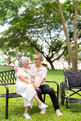 Fototapeta premium Elderly couple resting on a bench in the park with wheelchair is parked nearby.