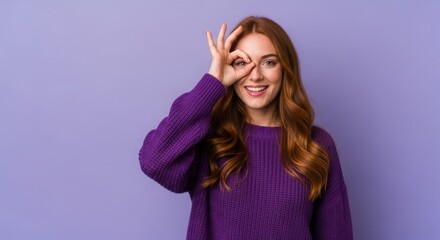 Young woman in a purple sweater playfully making an 'OK' gesture with her hand against a pastel background