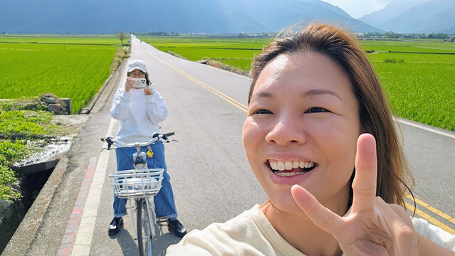 A young lesbian couple smiling cycling through Mr. Brown Avenue in Chishang, Taitung, Taiwan &mdash; romantic self-guided journey through rice paddies and countryside, backpacking travel selfie