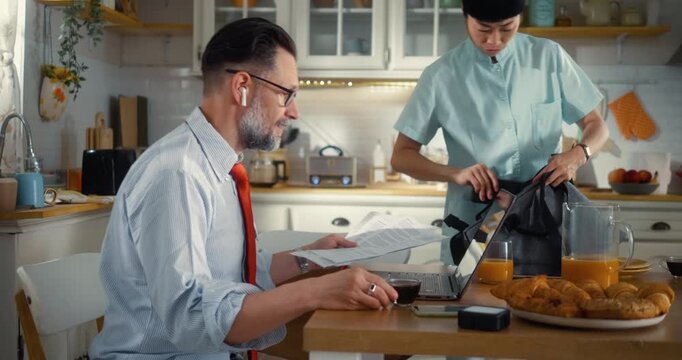 Remote working man in shirt and tie has video conference call in kitchen while his Japanese wife nurse, packs for work and leaves after a kiss goodbye in the morning.