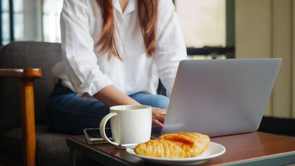 front view of woman using laptop computer, working from home in the morning with hot coffee, croissant bakery and mobile phone on the table. anonymous face