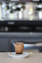 Close up of a delicious iced coffee drink with foam and chocolate shavings served in a clear glass on a white saucer in a coffee shop setting with a blurred background of a coffee machine and bokeh