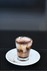 A Delicious Layered Coffee Drink with Cream and Cocoa Powder Served in a Small Glass on a White Saucer with a Dark Background