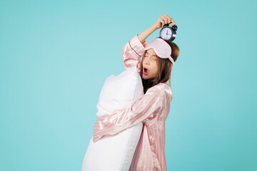 Tired woman in pajama with sleep mask embraces pillow, holding alarm clock above her head while...