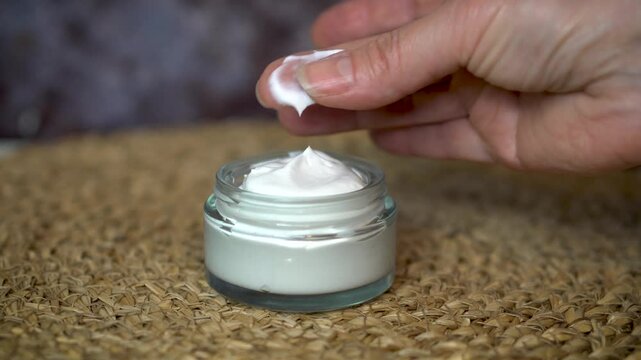 Woman's hand testing homemade natural face or body cream by rubbing it between fingers. Organic skincare in glass jar on beige and dark background. Clean beauty and eco-friendly concept.