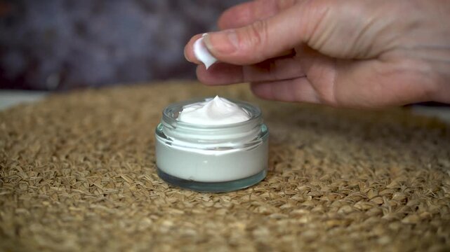 Woman's hand testing homemade natural face or body cream by rubbing it between fingers. Organic skincare in glass jar on beige and dark background. Clean beauty and eco-friendly concept.