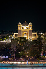 Fototapeta premium A beautifully lit cathedral or historic building stands tall at night in the heart of the city, surrounded by trees and a lively public square. Captured from above with a vibrant urban ambiance.