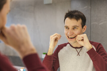 Young man maintains oral hygiene by flossing his teeth in front of the mirror, smiling and...