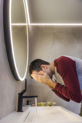 Young man refreshes his face with water in a modern bathroom, practicing daily facial hygiene and...