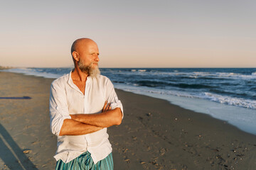 Confident older man with a white beard and linen shirt standing with arms crossed, gazing thoughtfully towards the sea. Shot at golden hour on a peaceful beach, conveying calm and introspection.