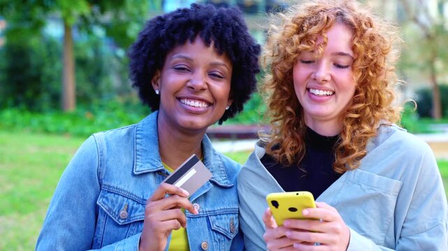  Cheerful African American and Caucasian friends stand in a city park, holding colorful smartphones and a credit card, celebrating cashless convenience, shared shopping success, and digital lifestyle