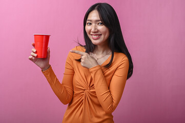 Asian woman with long black hair in orange dress smiling while pointing at a red plastic cup she is holding, standing against pink background, suggesting beverage promotion or party vibe