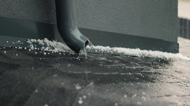 Rainwater flows from a gray downspout onto a roof covered with durable bitumen shingles during a heavy rainstorm with hailstones scattered around
