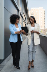 Two multiethnic businesswomen walking outside office discussing work while looking at digital tablet