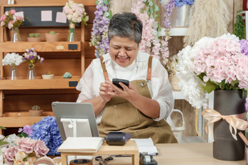 Asian elderly florist reading online order on mobile phone with joyful expression while seated at flower shop counter surrounded by artificial flower bouquets in creative working space