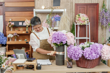 Asian senior florist decorating hydrangea bouquet with pink ribbon on wooden table in small indoor flower store while preparing arrangement for customer order in her home business after retirement