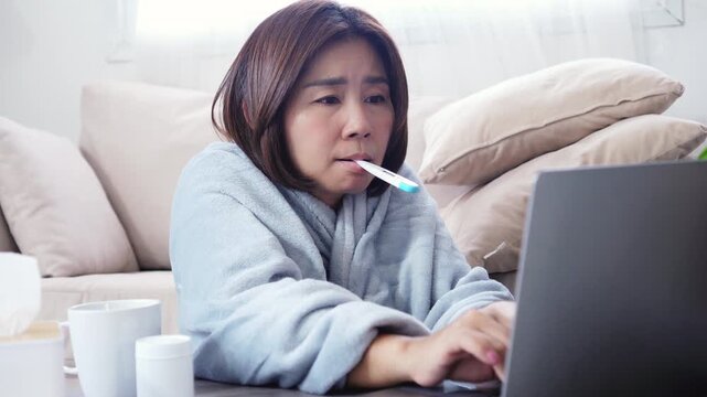 asian woman feeling very sick at home with high fever, using thermometer and resting under a blanket from cold or flu working online from home