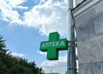 A green pharmacy cross with Cyrillic script "Аптека" (Pharmacy) hangs on a building against a blue sky.
Concept of Healthcare access.