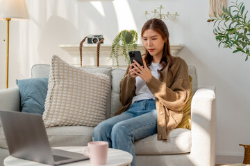 young woman sits on cozy sofa, using her smartphone while relaxing in bright living room. space...