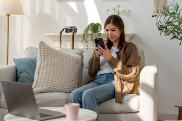 young woman sits on cozy sofa, using her smartphone in bright living room. space features plants,...