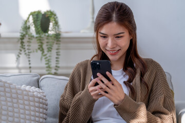 young woman smiles while using smartphone, sitting comfortably on sofa in cozy living room. natural...