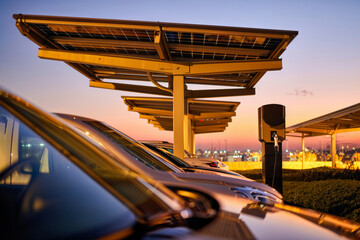 EV charging at sunset. An electric car charging at a station under a solar panel carport with a vibrant city sunset in the background.