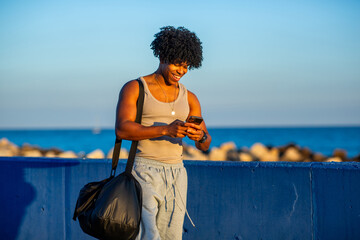 Smiling athletic mixed race man standing by the sea using smartphone and carrying sports bag