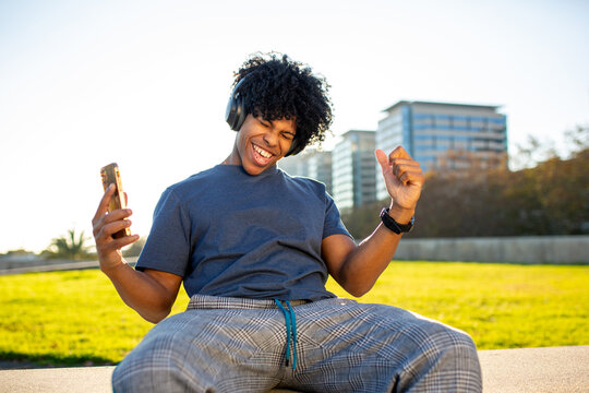 Mixed race young man dancing joyfully with headphones and smartphone in sunny park