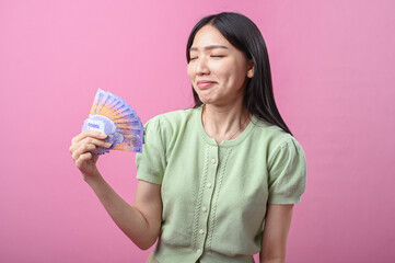 Asian woman in green top smiling confidently while holding a fan of purple Indonesian banknotes in one hand, standing against a solid pink background