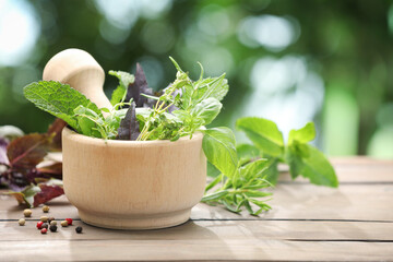 Mortar with different fresh herbs and pestle on wooden table against blurred green background, closeup
